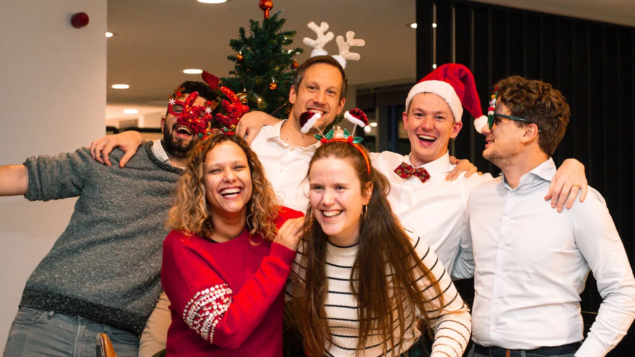 Six people in festive attire, wearing Christmas-themed accessories, smiling and posing in front of a decorated tree indoors.