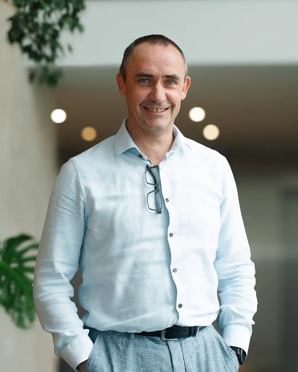 Smiling man in a light shirt with glasses, standing indoors with blurred background and plants.