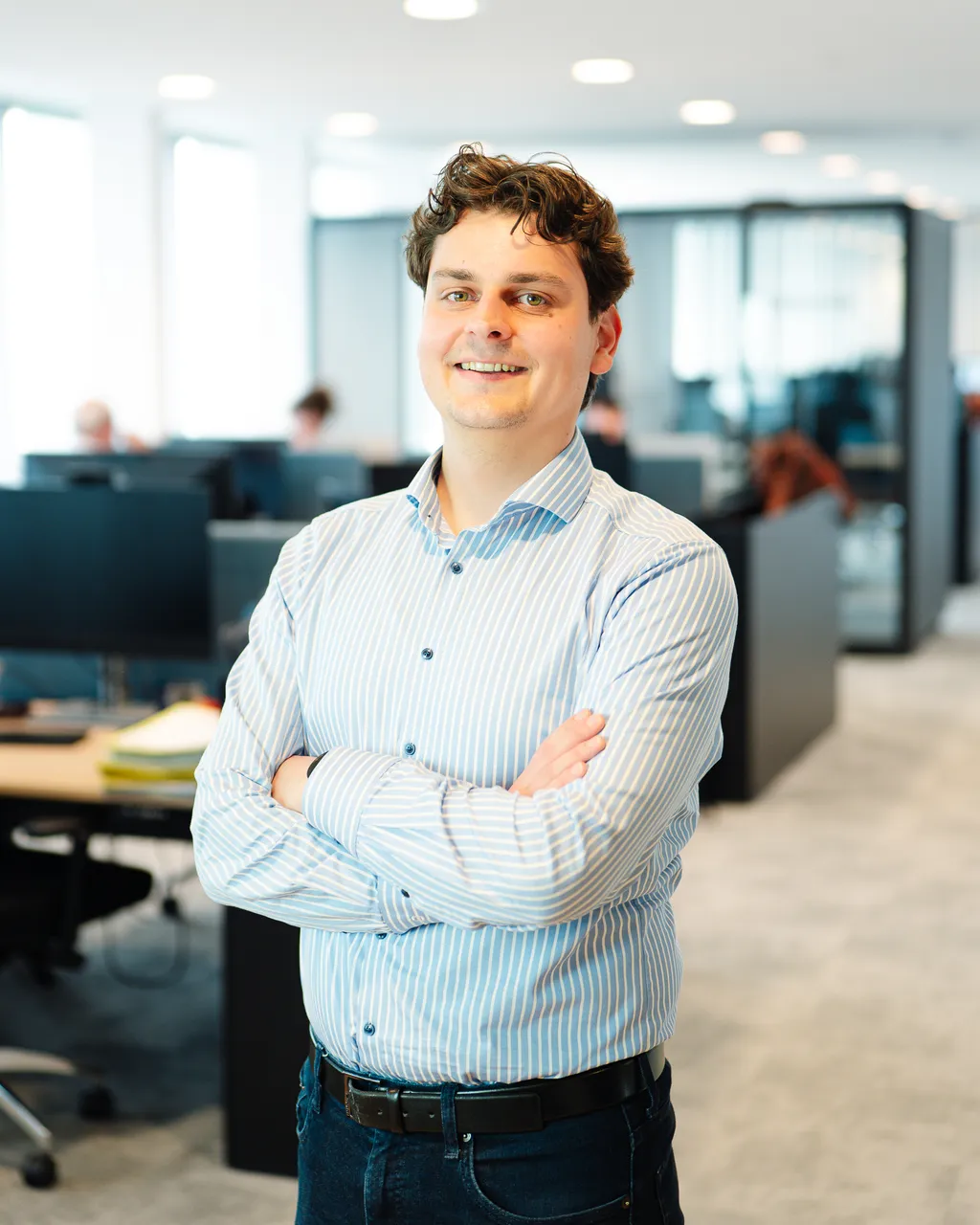 Smiling office worker in a striped shirt stands with folded arms in a bright, modern open-plan workspace.