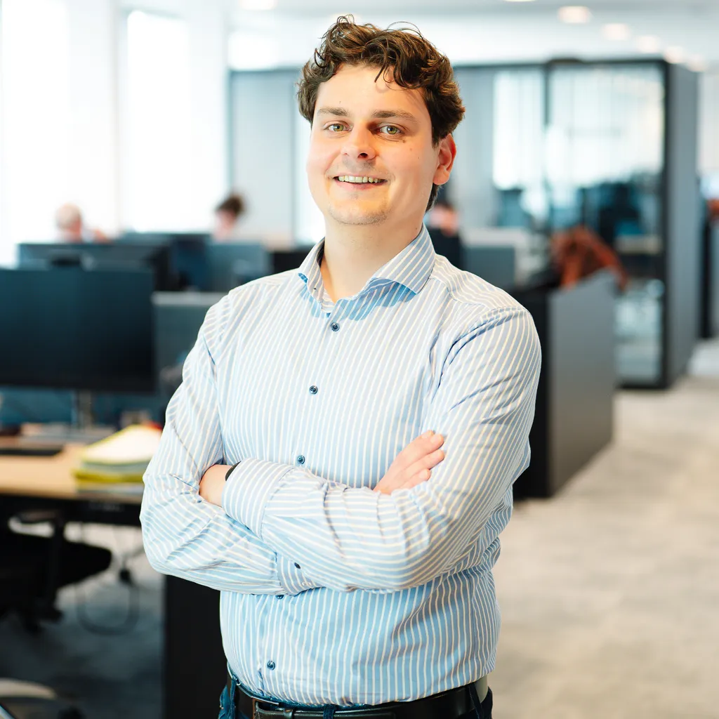 Smiling office worker in a striped shirt stands with folded arms in a bright, modern open-plan workspace.