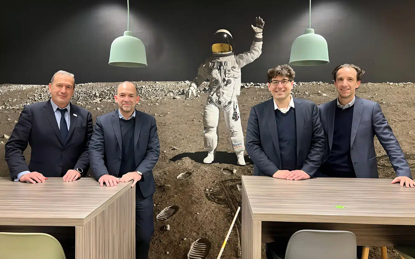Four men in suits stand at a table, with an astronaut mural prominently displayed on the wall behind them.
