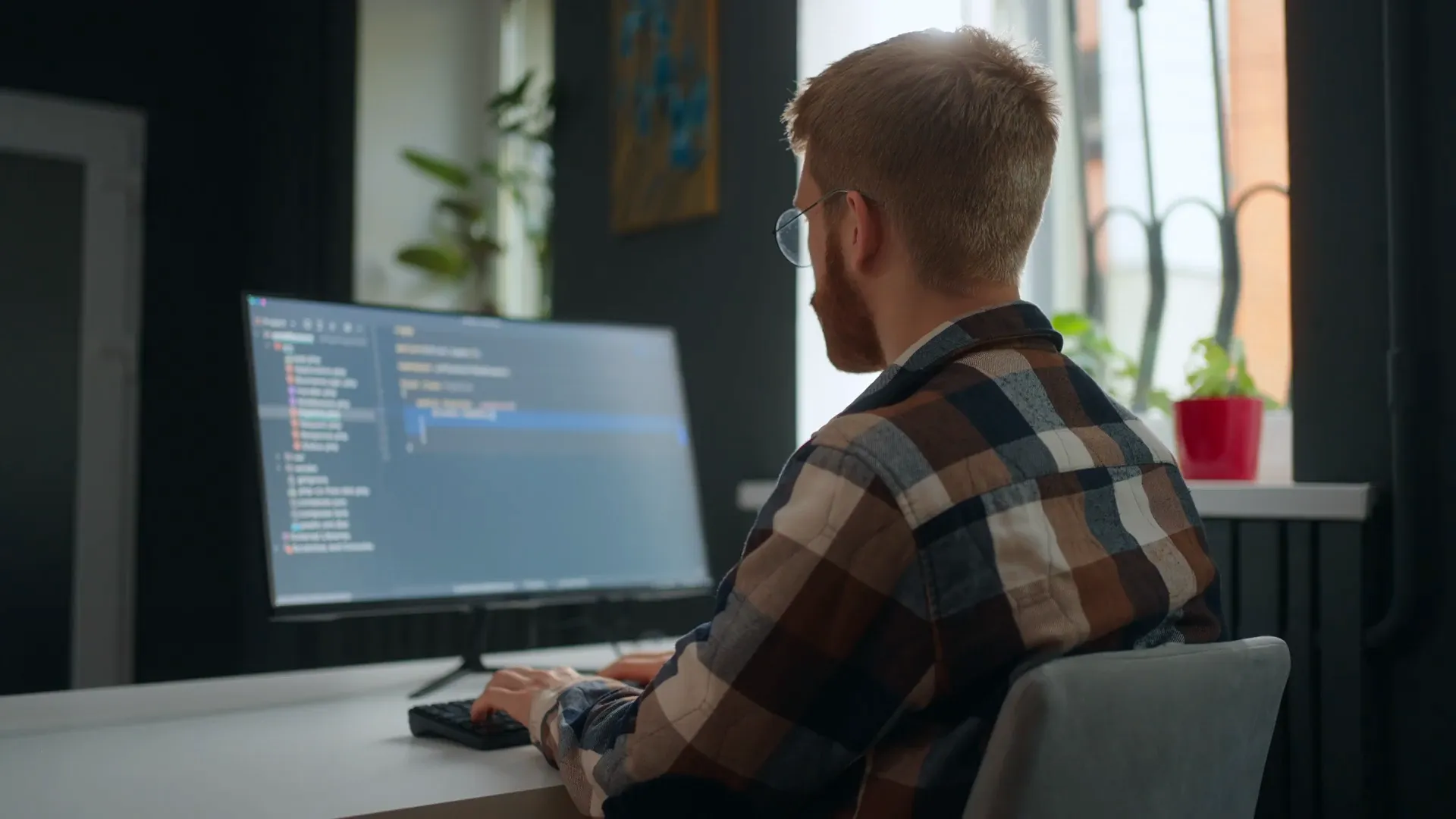 A man in a plaid shirt sits at a desk, focused on his computer screen, surrounded by a tidy workspace.