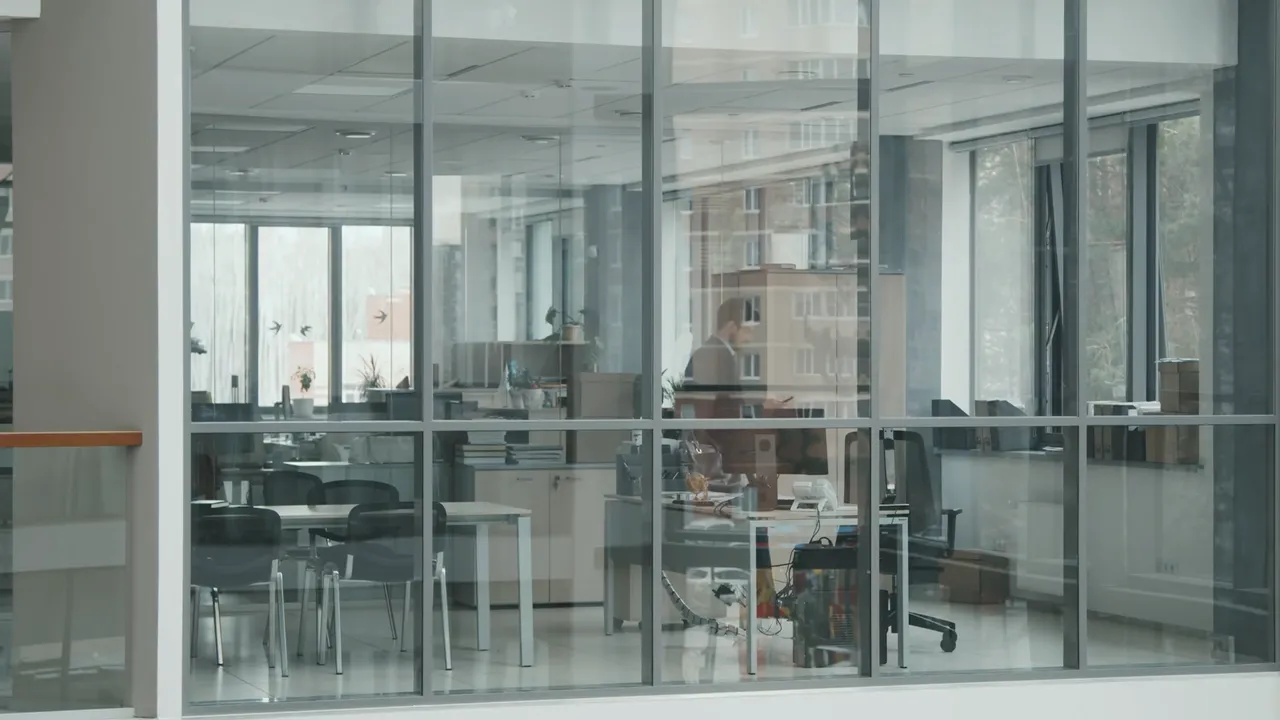 Male colleague looking at his phone while walking in his desk. Shot from outside through the window.