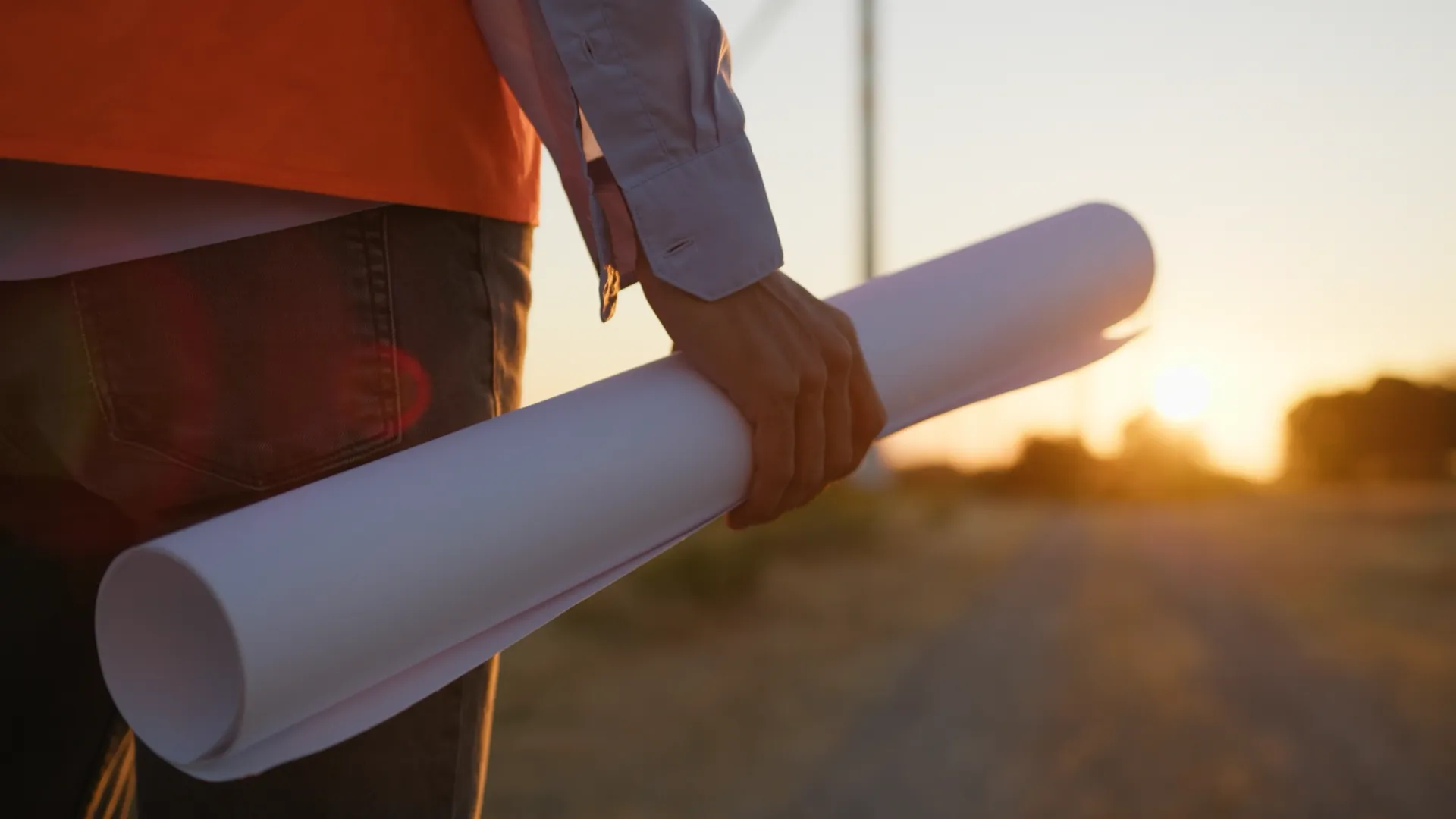 Persoon houdt opgerolde bouwtekeningen vast terwijl hij over een bouwterrein loopt tijdens zonsondergang.