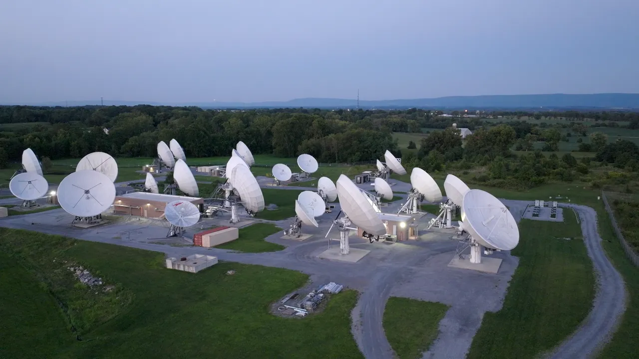 Aerial view of a satellite communication facility with multiple large white parabolic satellite dishes arranged across a green landscape. The facility includes small buildings and equipment, with a gravel road winding through the area. The background features rolling hills, trees, and a soft blue evening sky.