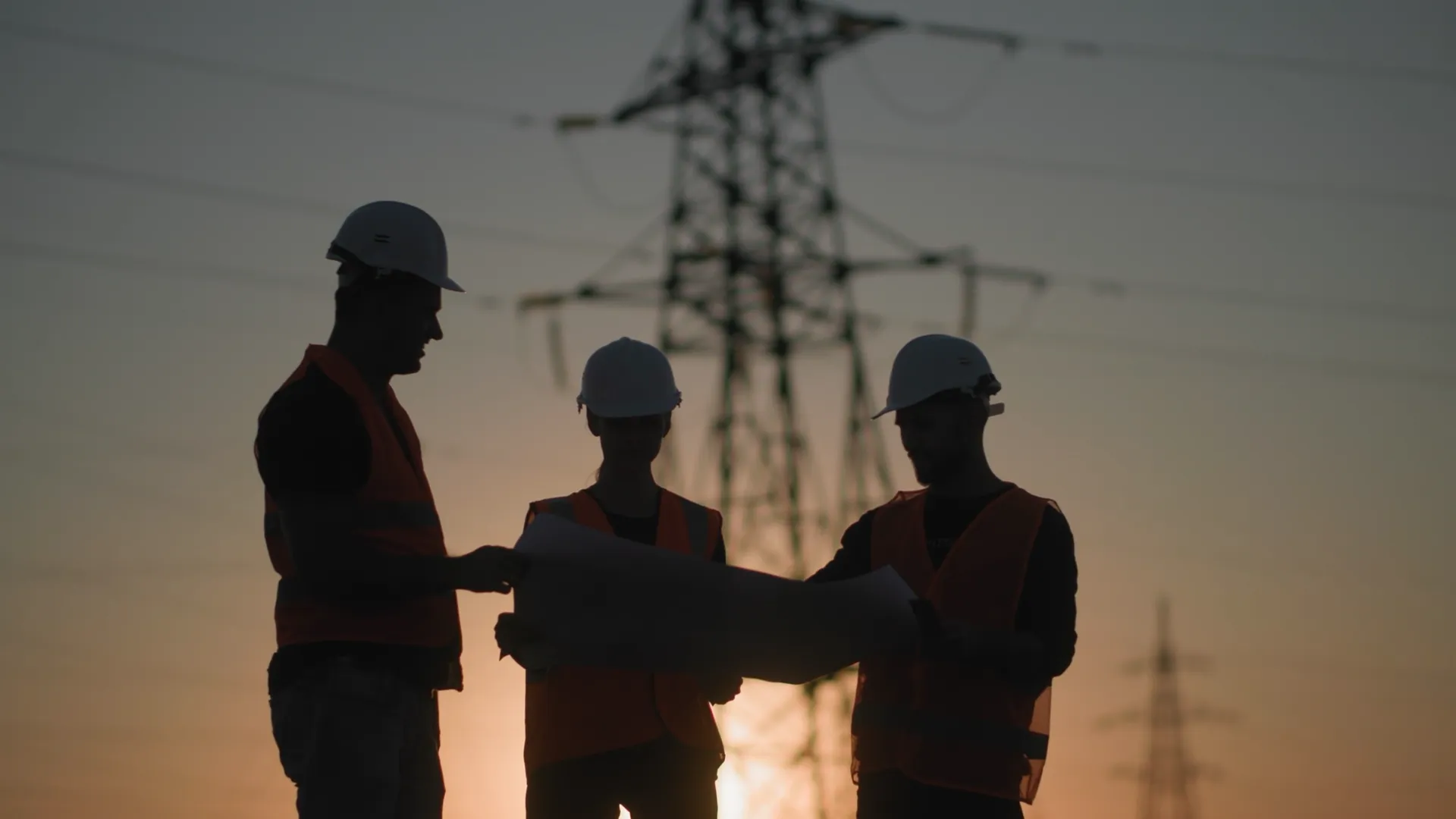Drie engineers in veiligheidskleding bekijken bouwplannen bij een hoogspanningsmast tijdens zonsondergang.