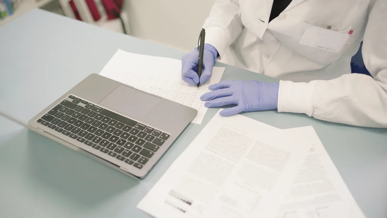 A person in a lab coat writing notes on a piece of paper at a laboratory desk.