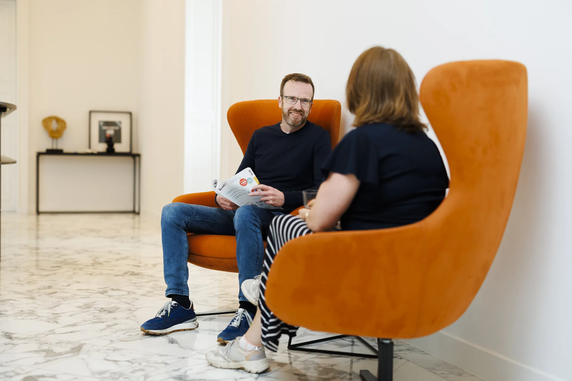 Man sitting in a chair over another woman seemingly in a conversation