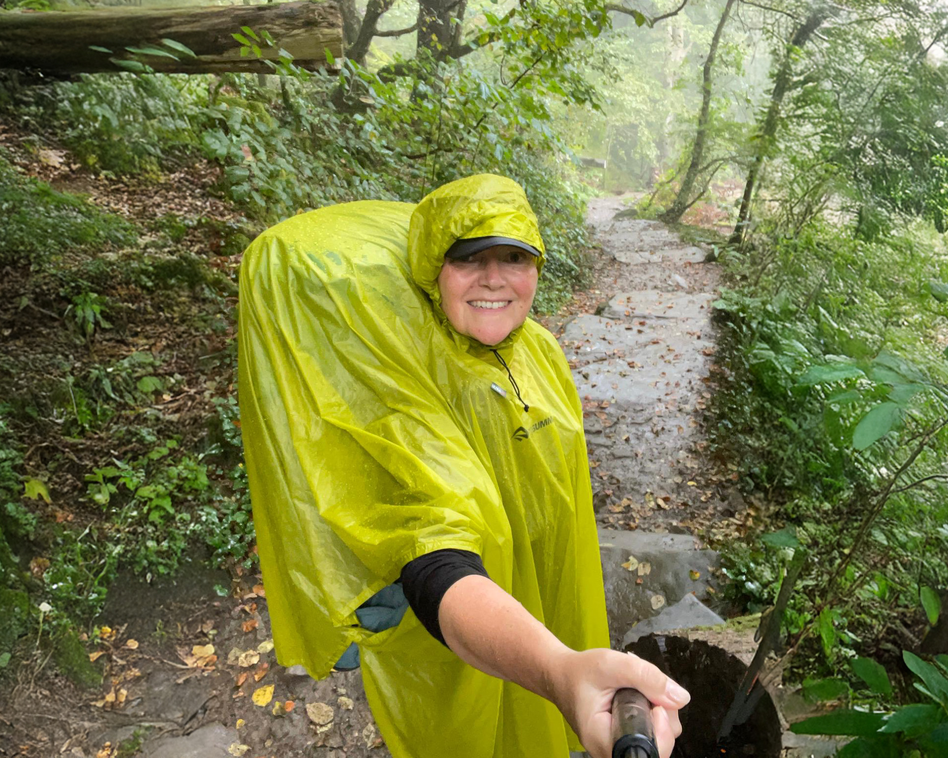 Person in a yellow rain poncho hiking on a forest trail, smiling and holding a selfie stick, surrounded by lush greenery and wet foliage.