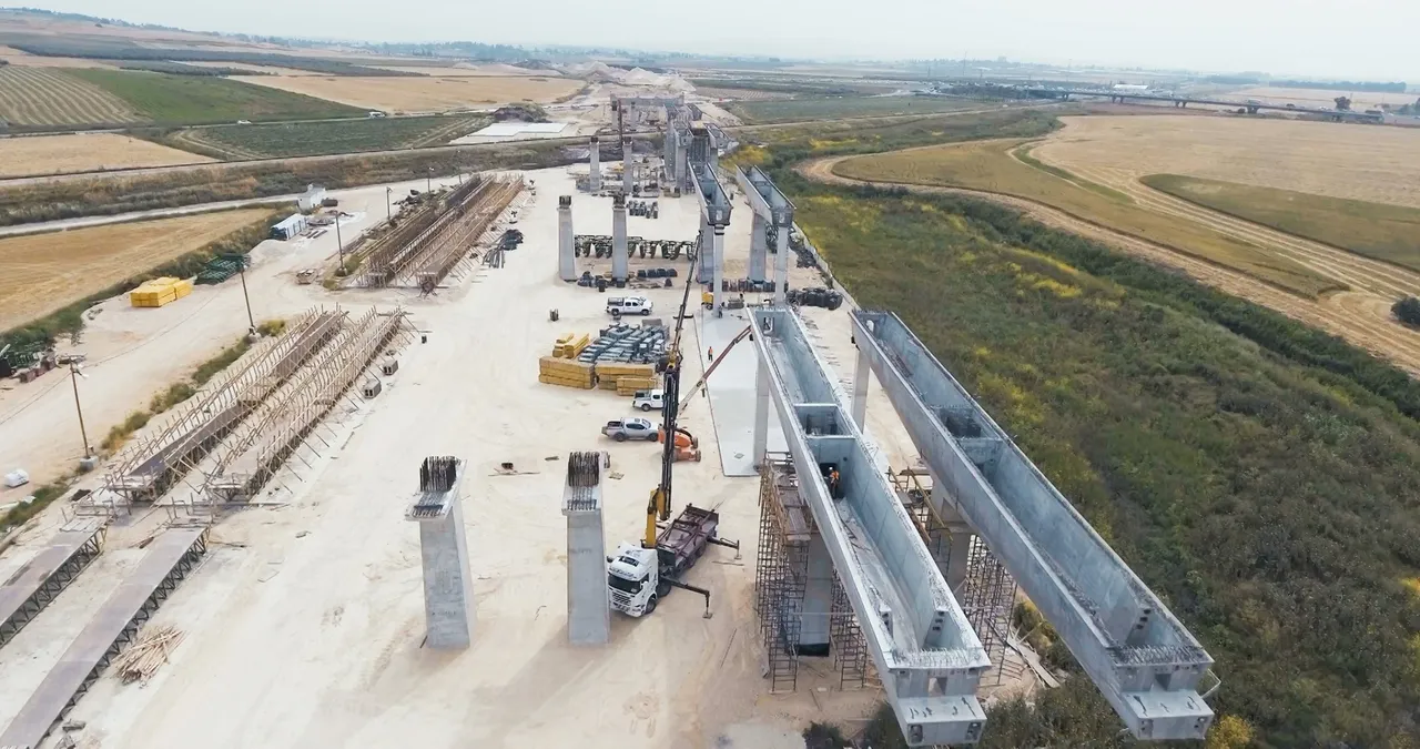 Aerial view of a highway bridge under construction, with concrete pillars, cranes, and materials on a sandy construction site surrounded by fields.