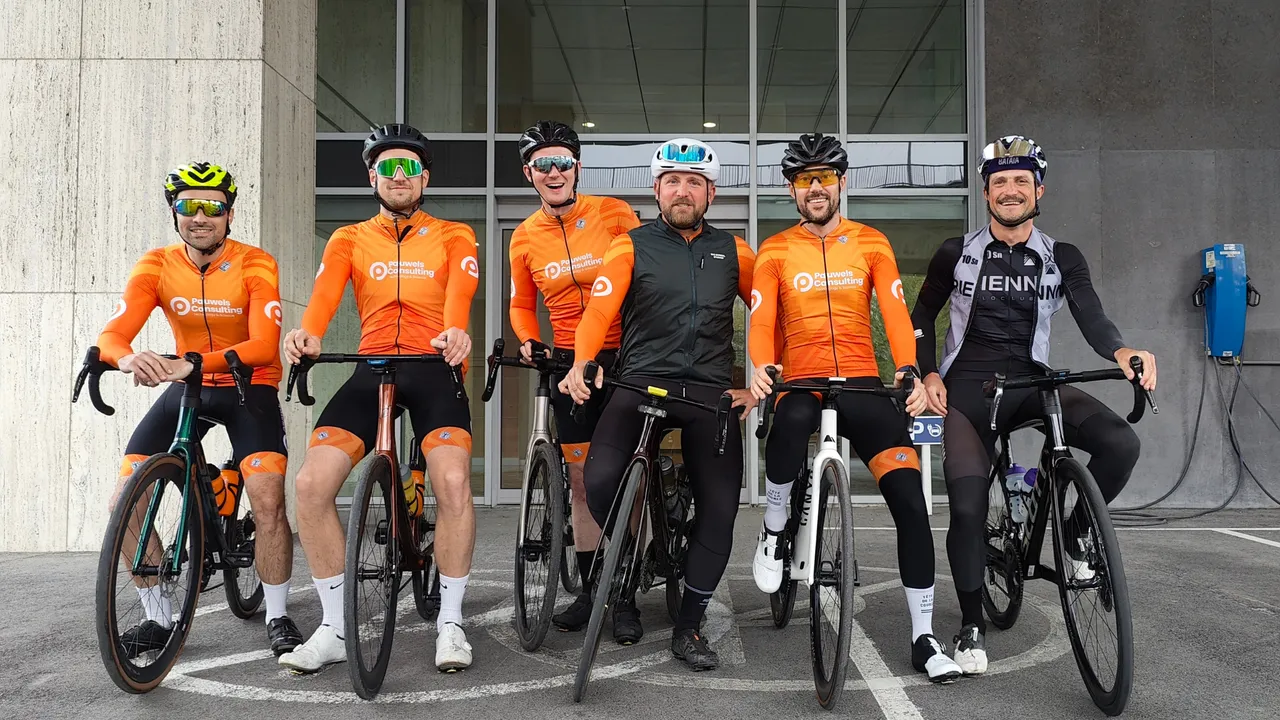 Six cyclists in bright orange jerseys and one in black, pose with their bikes in front of a modern building.