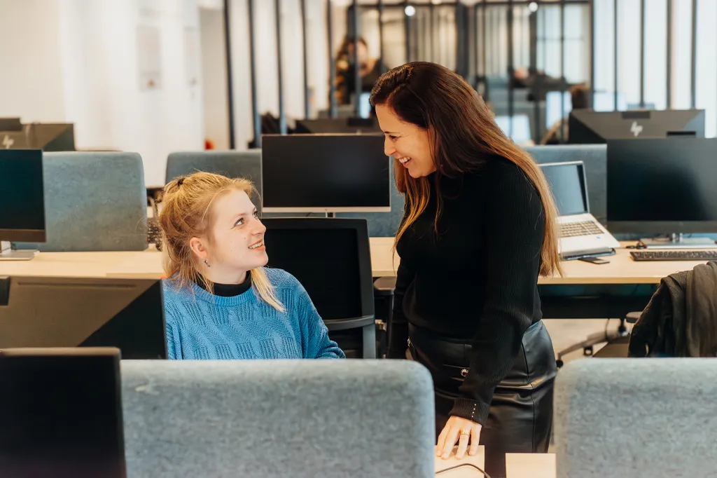 Two women interact in a modern office; one seated at a desk, the other standing and smiling. Computers and office cubicles surround them.