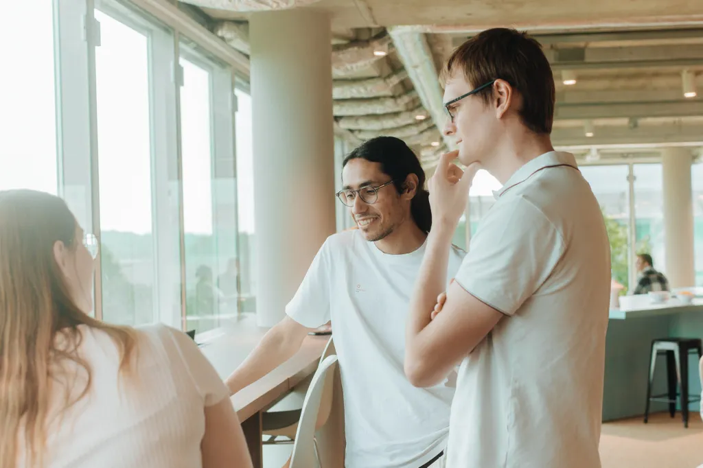Three people in casual attire have a conversation by large windows in a modern office space, with natural light streaming in.