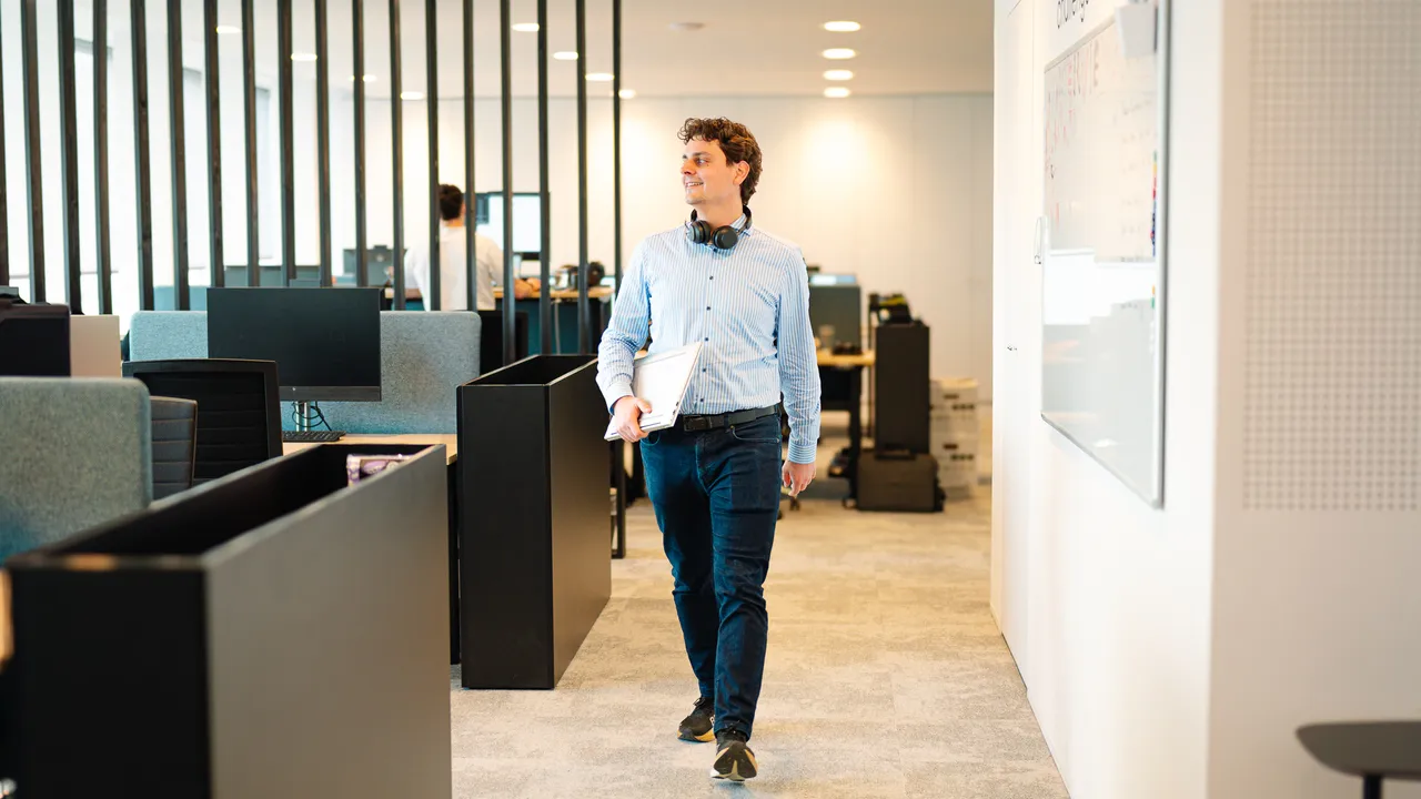 Man in business casual clothes walking through modern open-plan office, holding a laptop and wearing headphones around his neck.