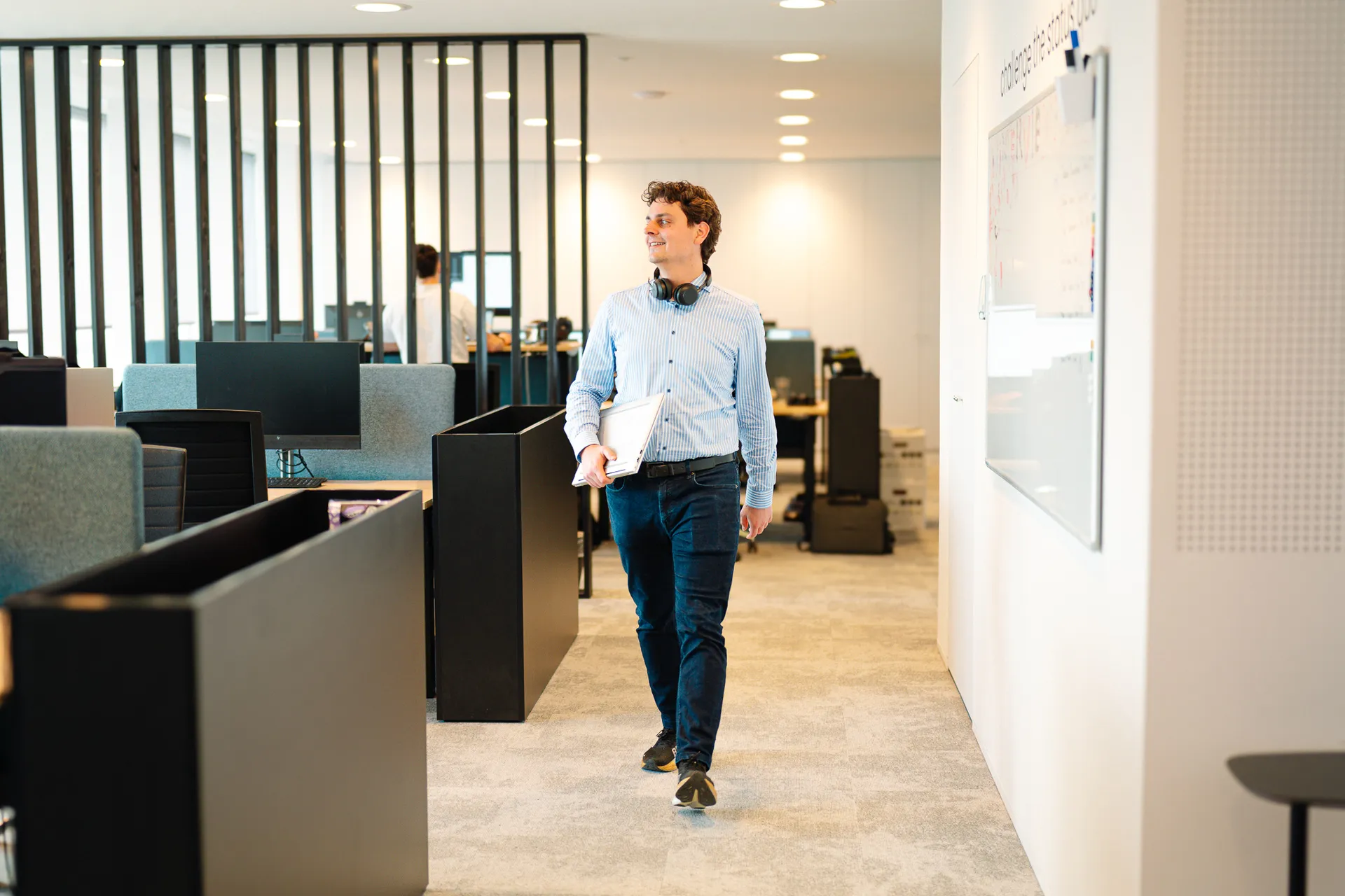Man in business casual clothes walking through modern open-plan office, holding a laptop and wearing headphones around his neck.