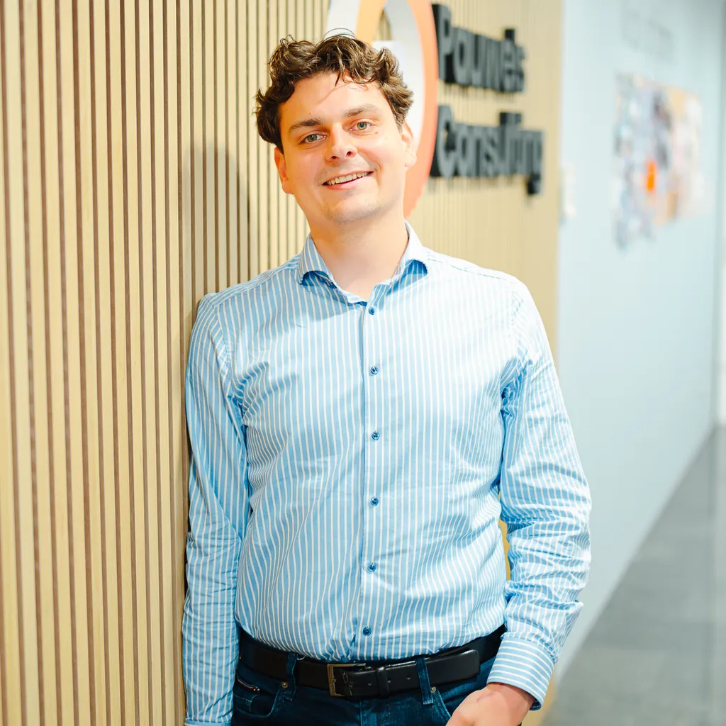 Smiling man in a light blue striped shirt leans against a wooden wall in a bright office hallway.