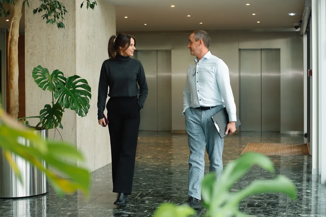 A woman and a man walking and talking in a modern office hallway with plants, wearing business attire, and holding a phone and a folder.