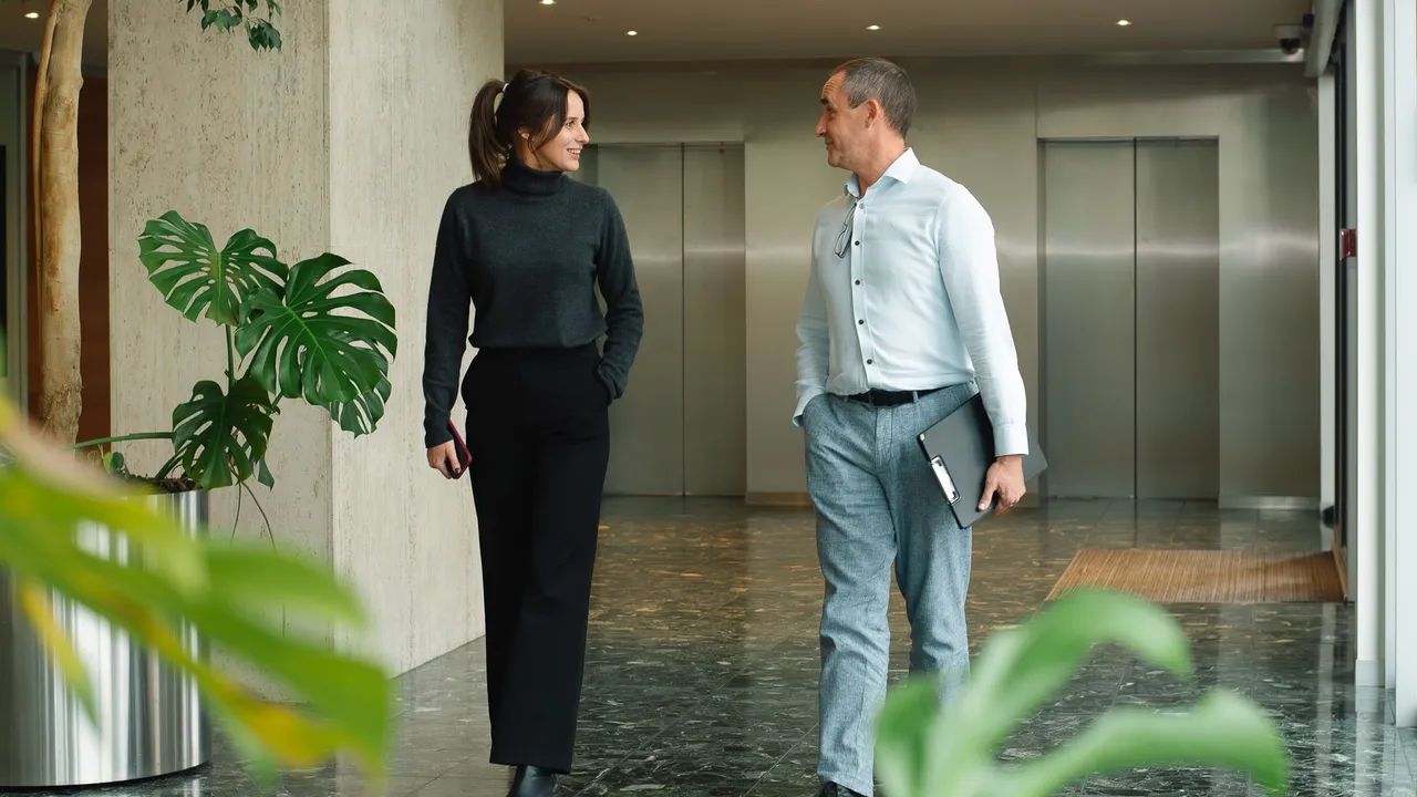 A woman and a man walking and talking in a modern office hallway with plants, wearing business attire, and holding a phone and a folder.