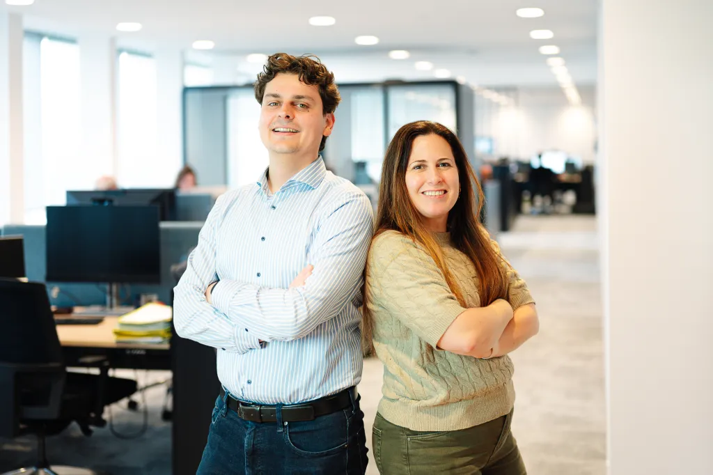 Two smiling coworkers standing back to back with arms crossed in a bright modern office.