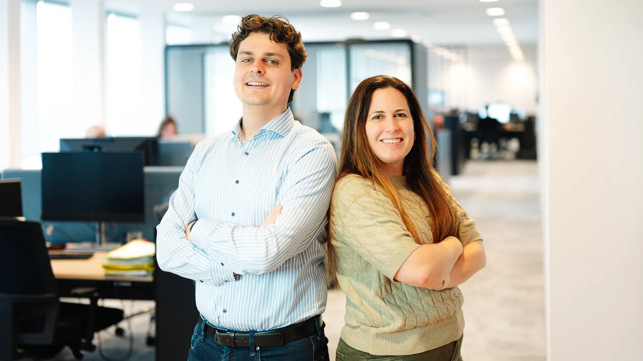 Two smiling coworkers standing back to back with arms crossed in a bright modern office.