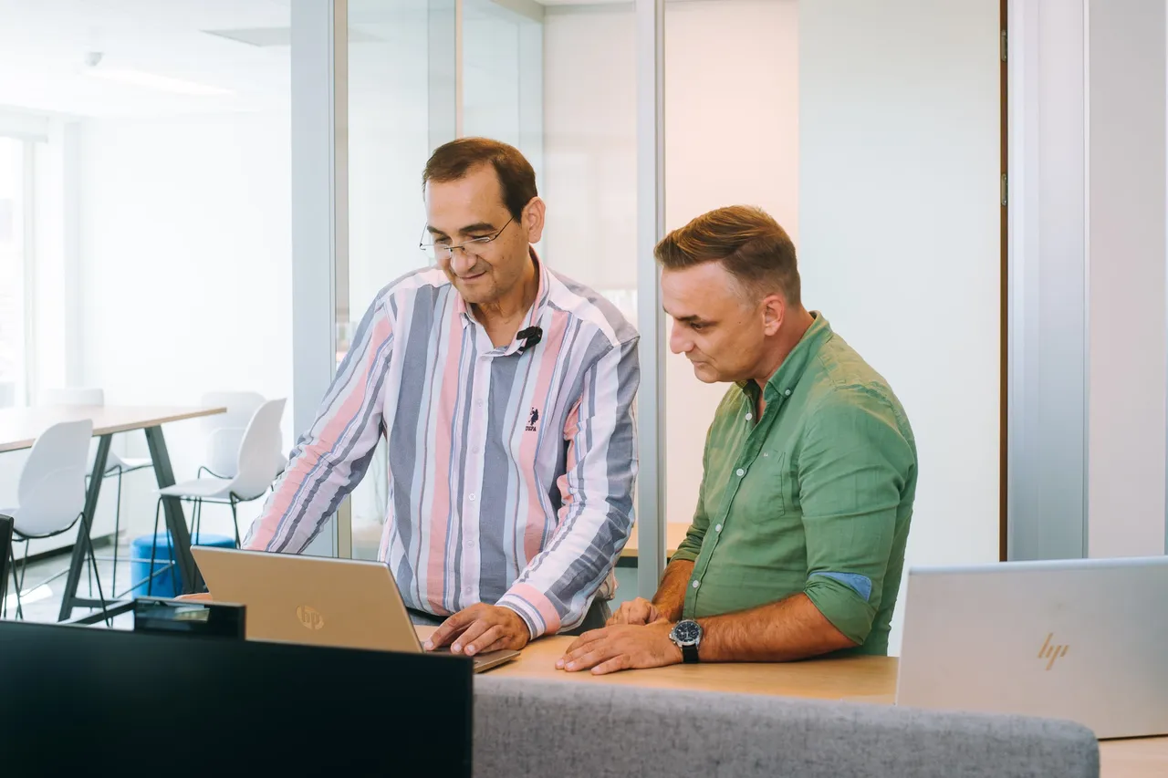 Two men stand at a desk in an office, looking at a laptop. One wears a striped shirt, the other a green shirt. Bright, modern setting.