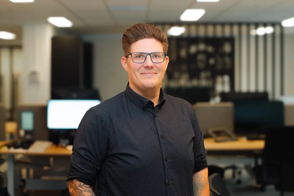 Person with glasses and tattoos, wearing a dark shirt, stands smiling in a modern office with computers and soft lighting in the background.