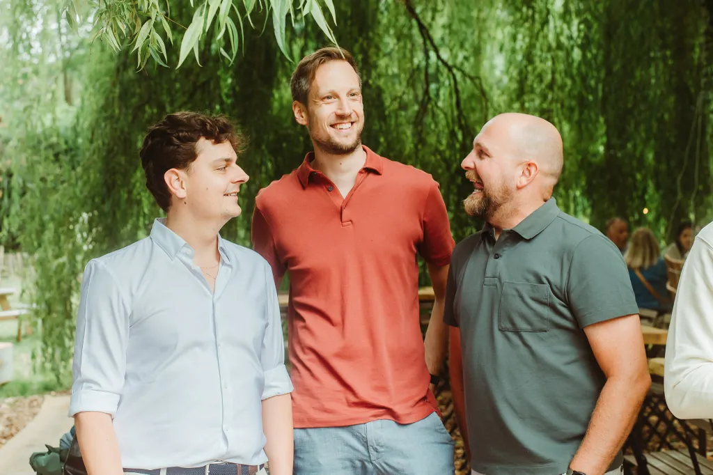 Three men smiling and chatting outdoors under lush green trees, with a relaxed and cheerful atmosphere.