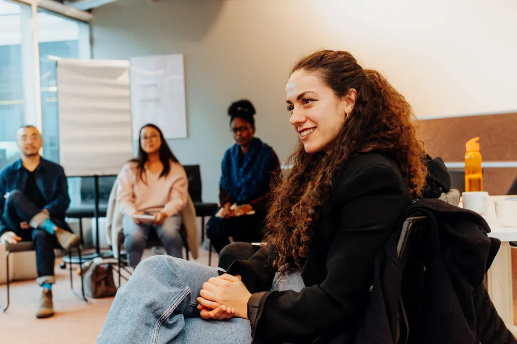 A woman with curly hair smiles while sitting in a group discussion. Others are seated in the background, with a flip chart visible.