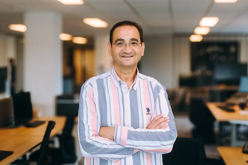 A man in a striped shirt stands confidently with arms crossed in a modern office setting.