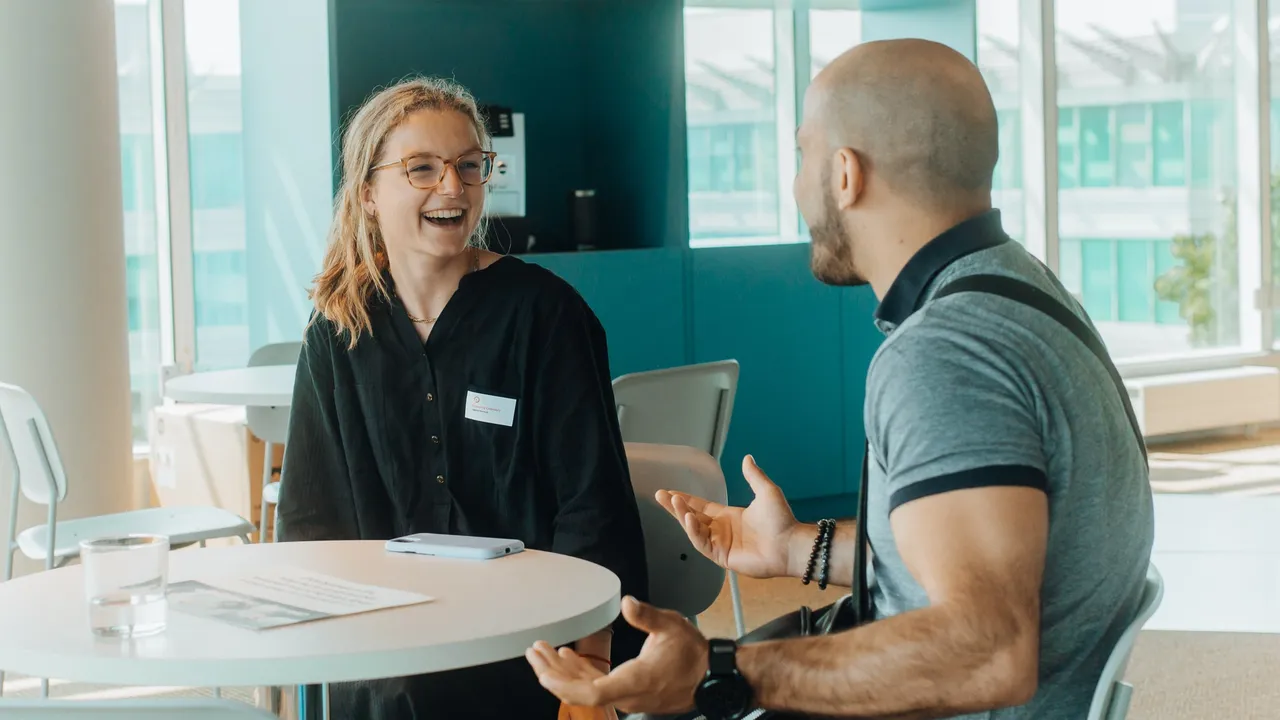 Two people sitting at a table in a bright room, engaged in a lively conversation. One is smiling, and the other gestures expressively.