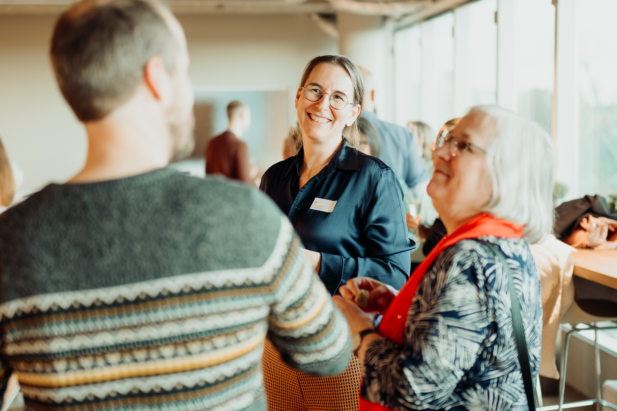Smiling woman with glasses chats with two people at a bright, casual networking event indoors.