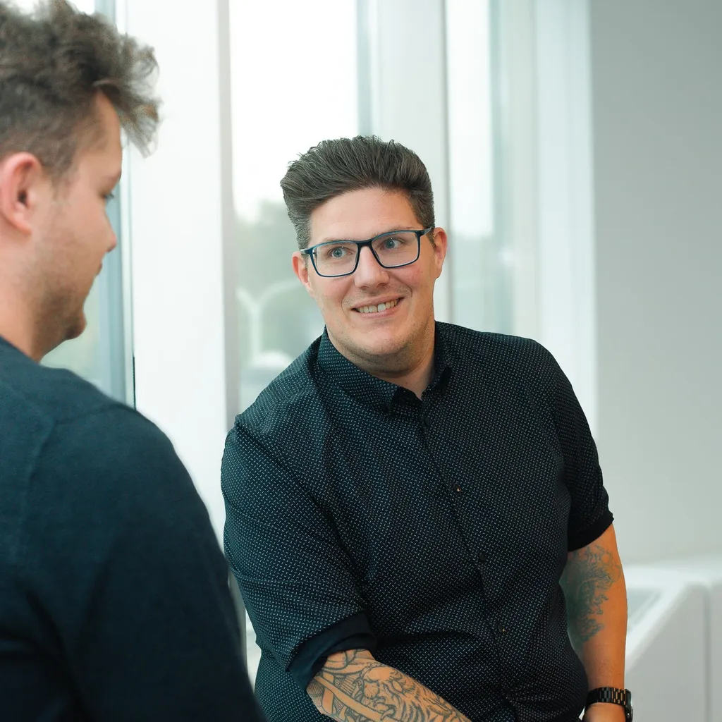 Two people talking by a window. One has glasses and tattoos, wearing a black shirt. The other has short hair and a dark sweater.