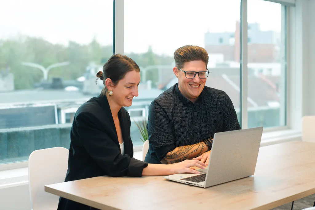 Two people smiling and looking at a laptop in a bright office with large windows, overlooking a cityscape.