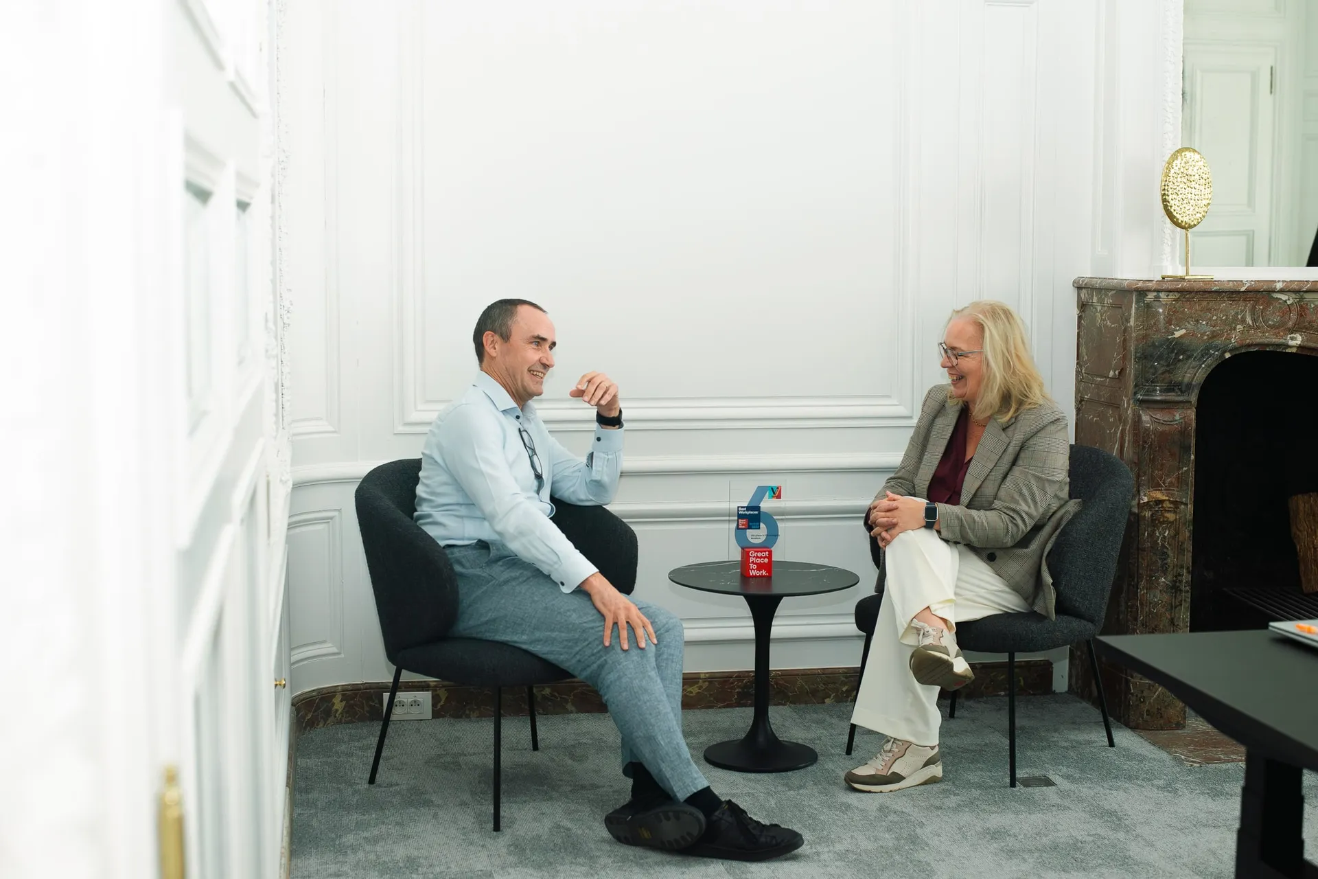 Two people sitting in a modern, white-walled room, engaged in conversation. A red soda can is on a small table between them.