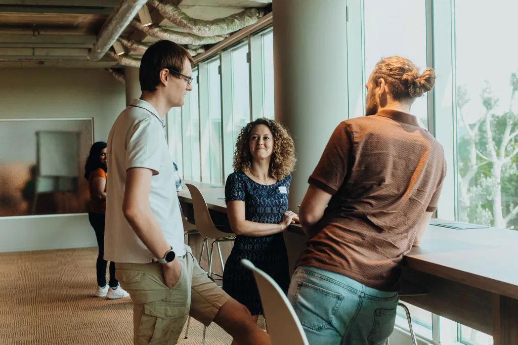 Three people casually chatting by a window in an office space, with one person leaning on a table. Two others are visible in the background.