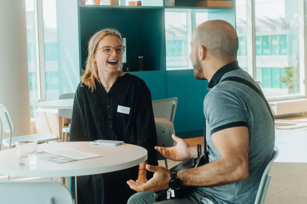 Two people sitting at a table in a modern office space, engaged in a lively conversation, both smiling and gesturing expressively.