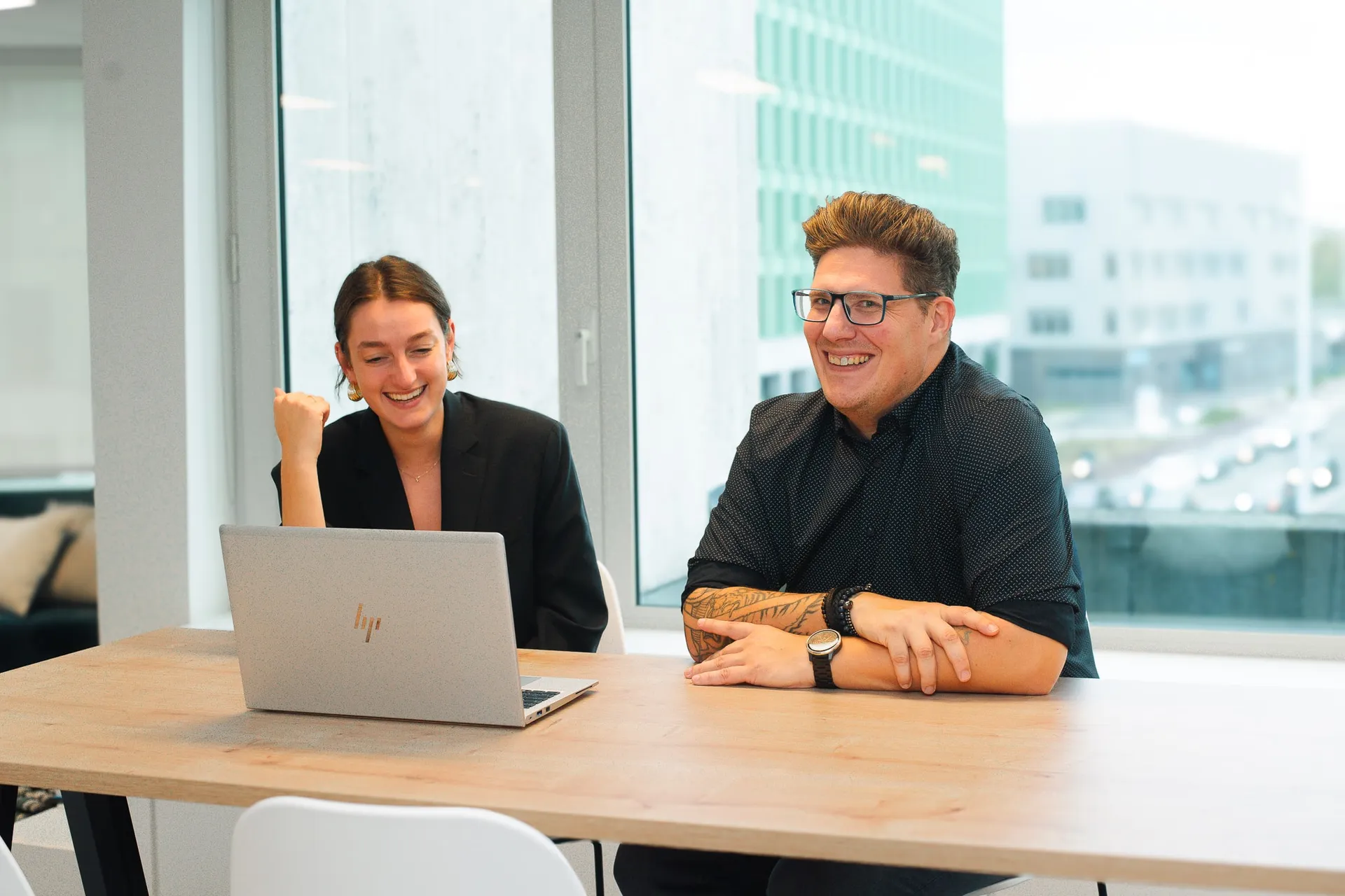 Two people sitting at a table with a laptop, smiling and engaged in conversation in a bright office setting.