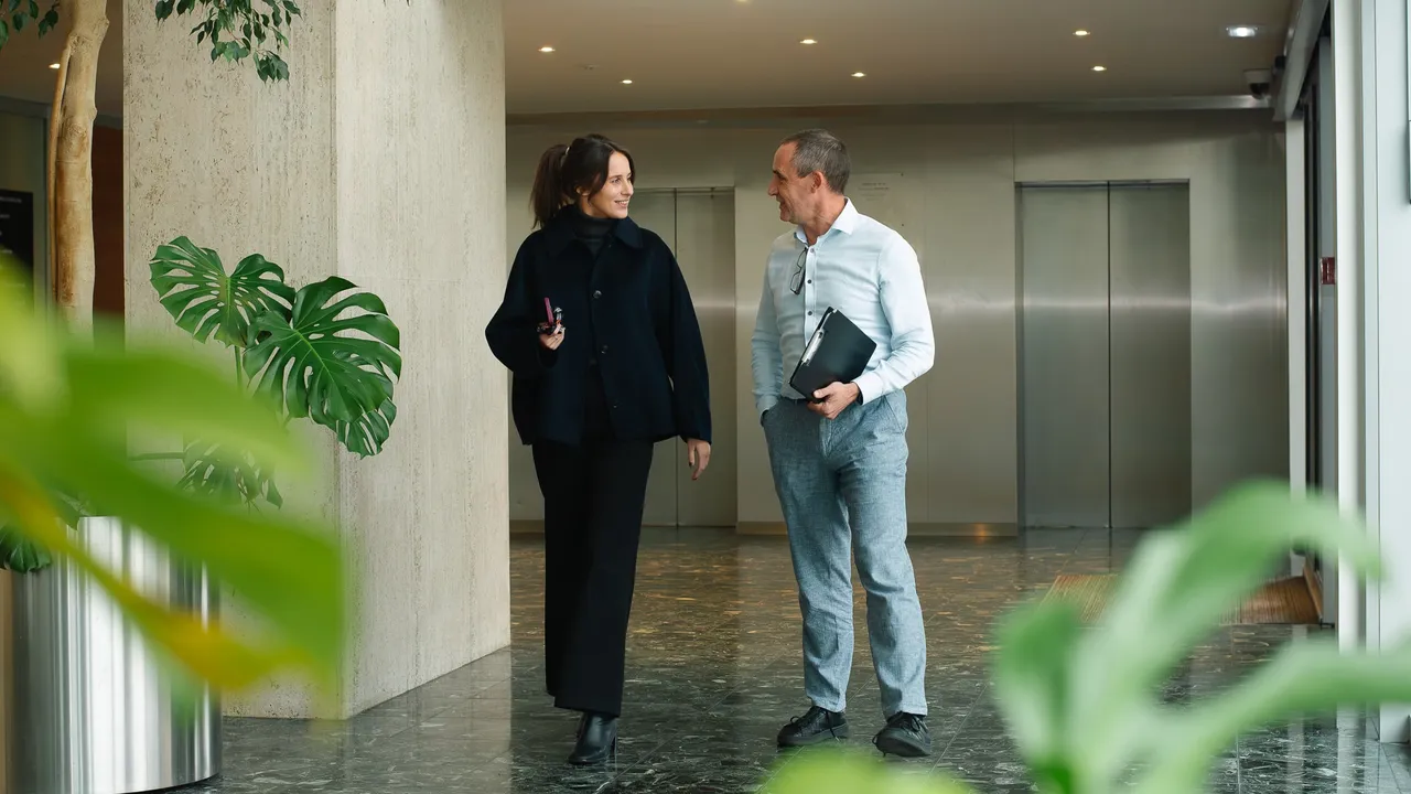 Two people walking and talking in a modern office lobby with plants and elevators in the background.