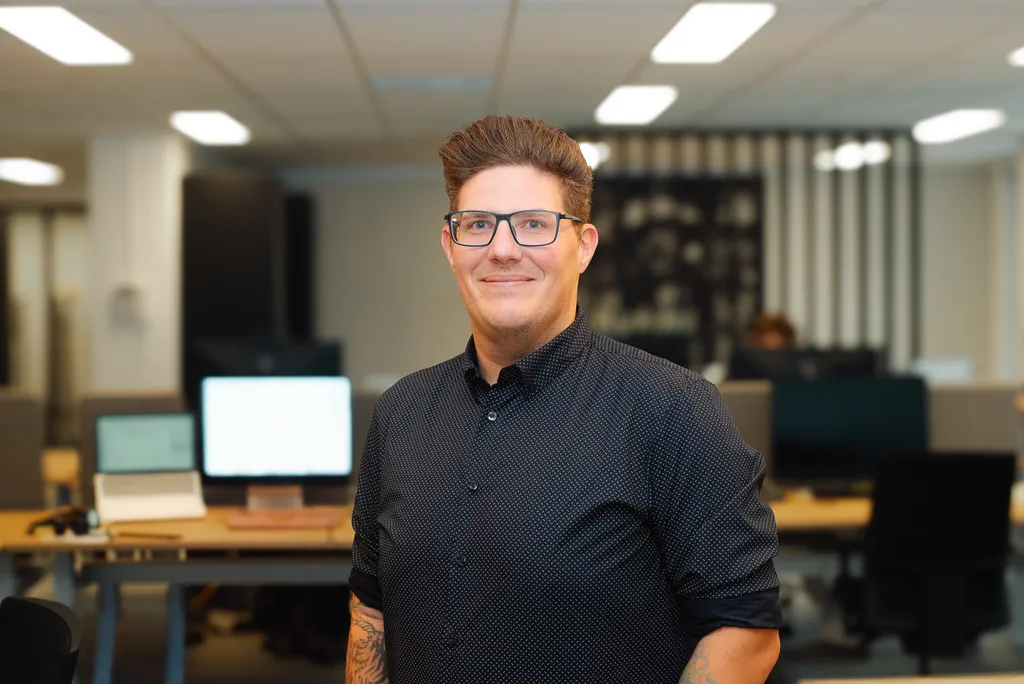 A person with glasses and tattoos, wearing a dark shirt, stands smiling in a modern office with computers and desks in the background.