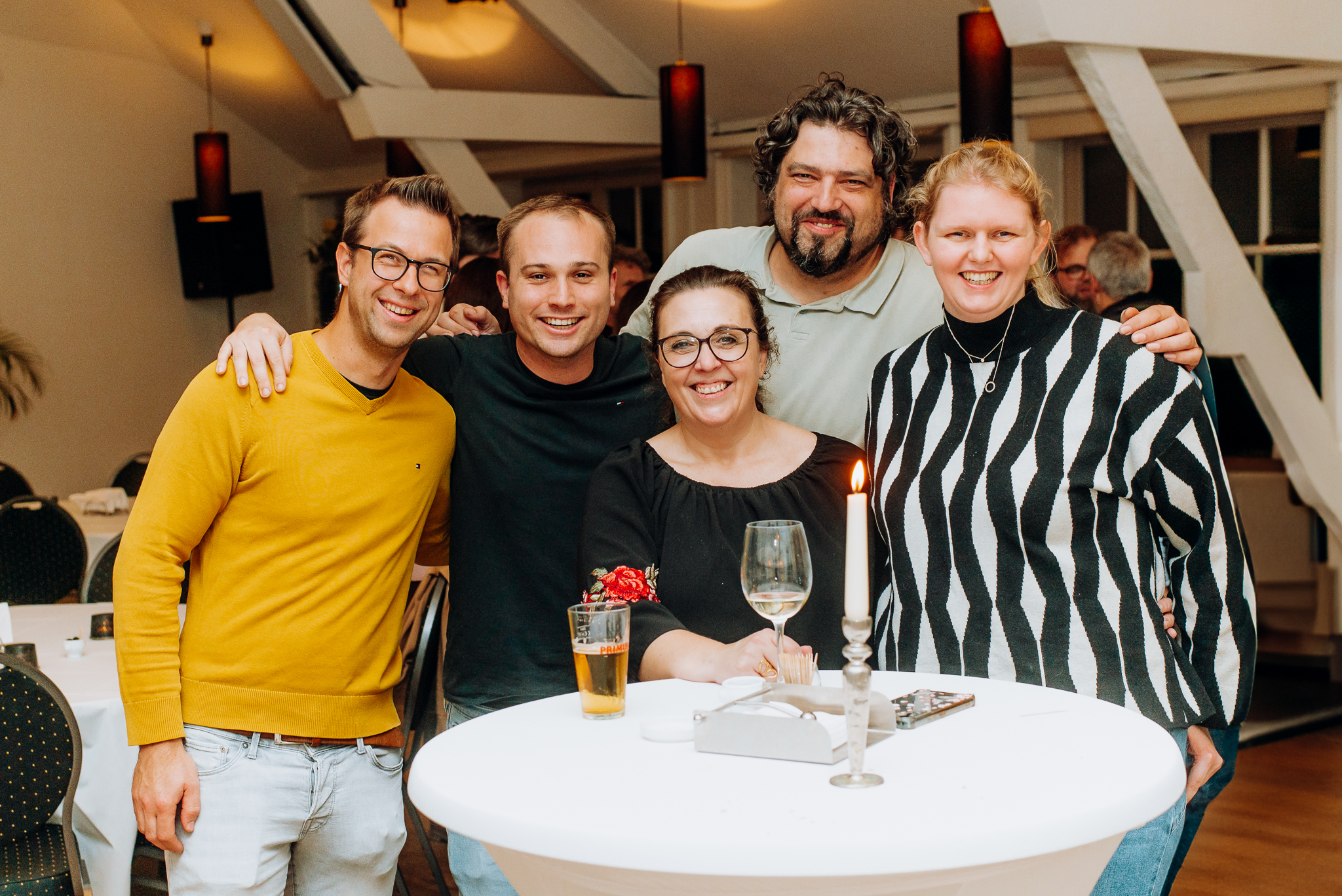 Five friends smiling and posing around a tall table with drinks and a lit candle at an indoor gathering.