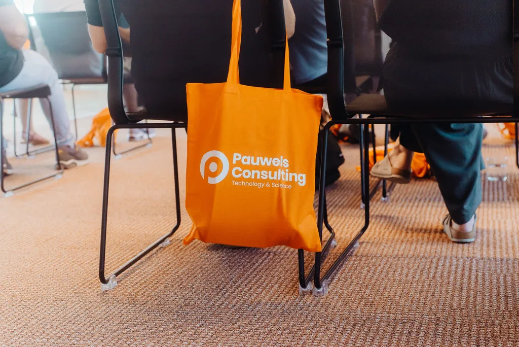 Orange tote bag with "Pauwels Consulting Technology & Science" logo hangs on a chair, surrounded by seated people on a carpeted floor.