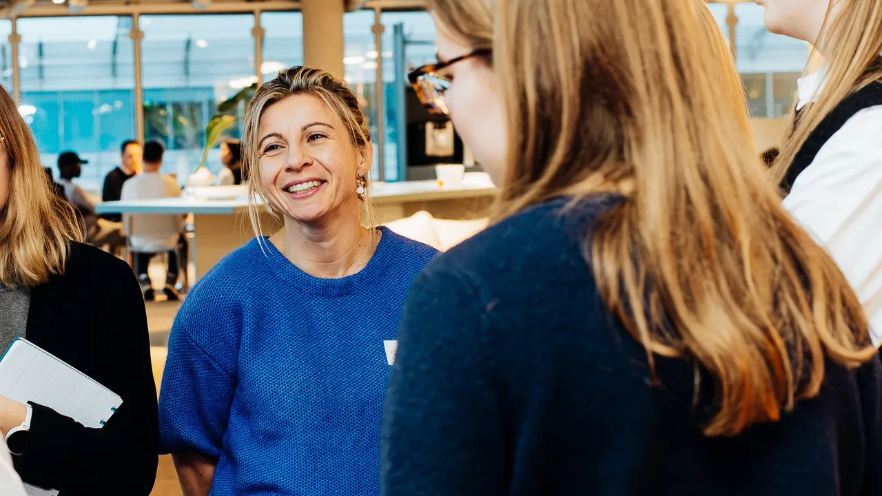 Smiling woman in a blue sweater talking with colleagues in an open-plan office, others blurred in foreground and background.