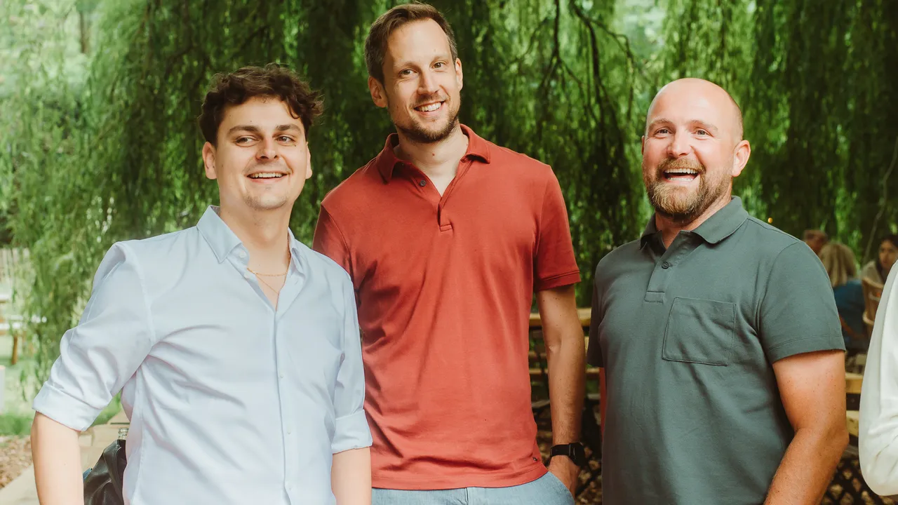 Three men smiling outdoors under lush green trees, wearing casual shirts.