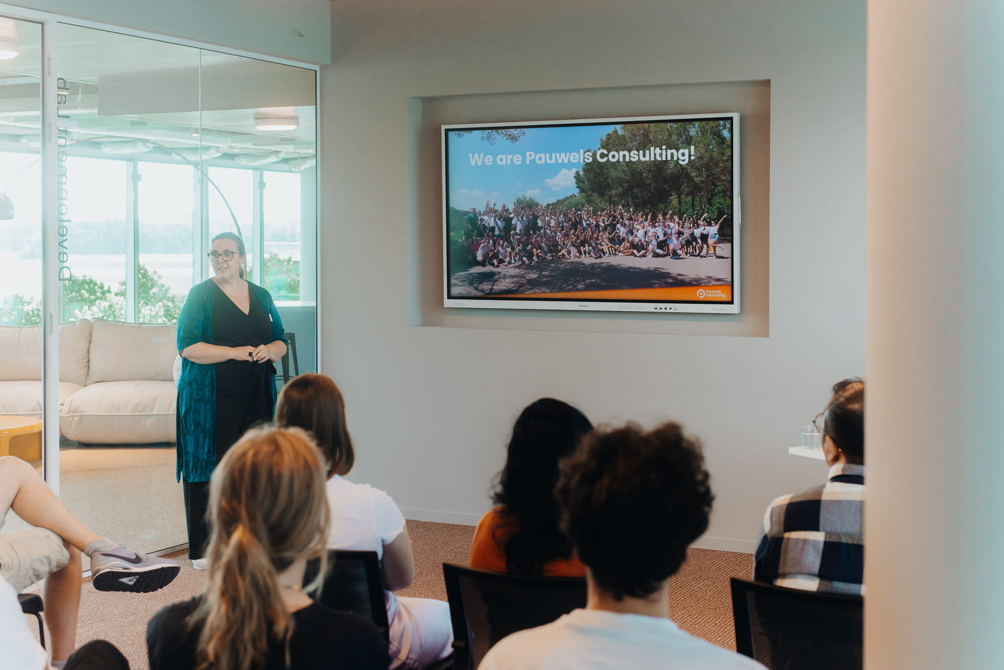 Woman giving a presentation to a seated audience, with a large screen showing “We are Pauwels Consulting!” and a team photo.