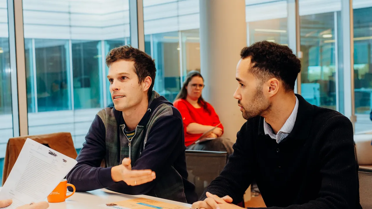 Two men engaged in discussion at a table, with a woman in red seated in the background. Papers and a mug are on the table.