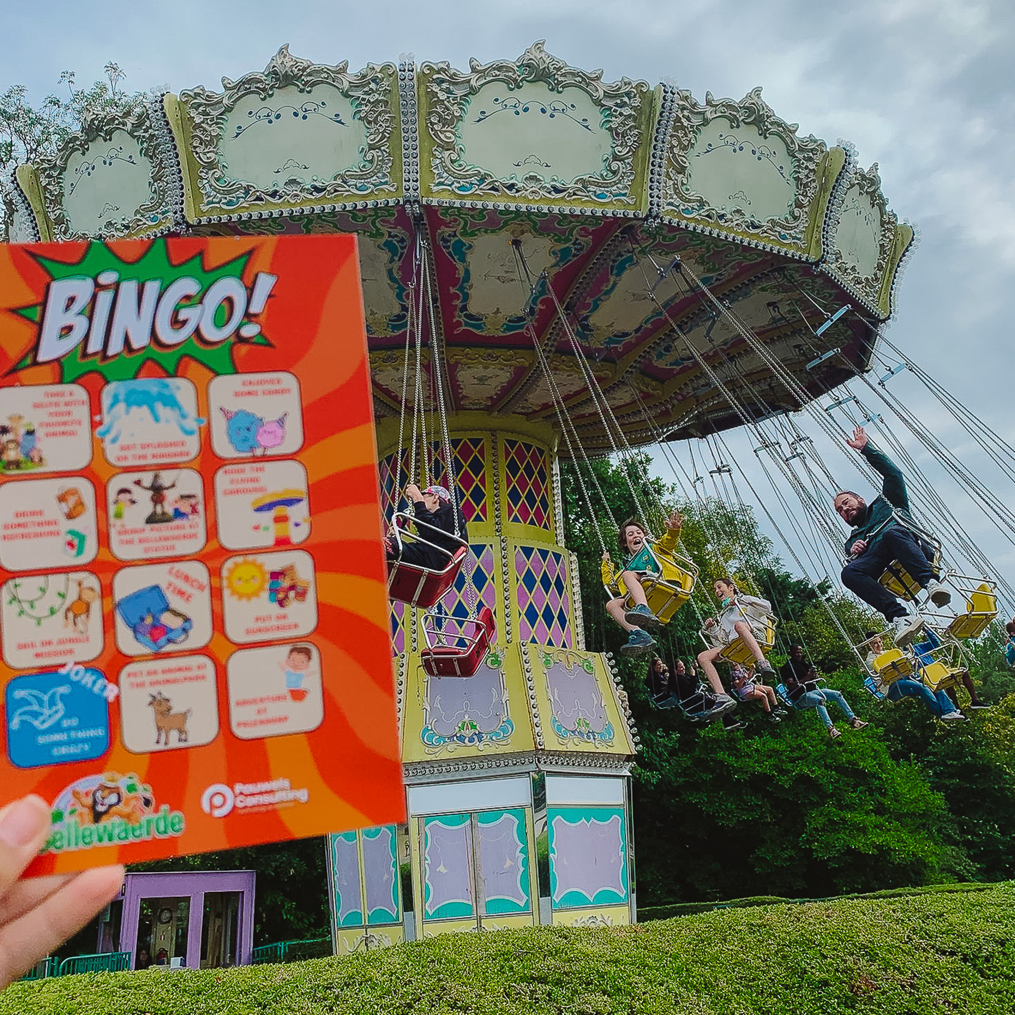 A colorful bingo card is held up in front of a spinning swing ride at an amusement park, with people waving and enjoying the ride.