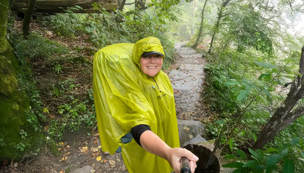 Person in a yellow rain poncho hiking on a misty forest trail, holding a selfie stick, surrounded by lush greenery.