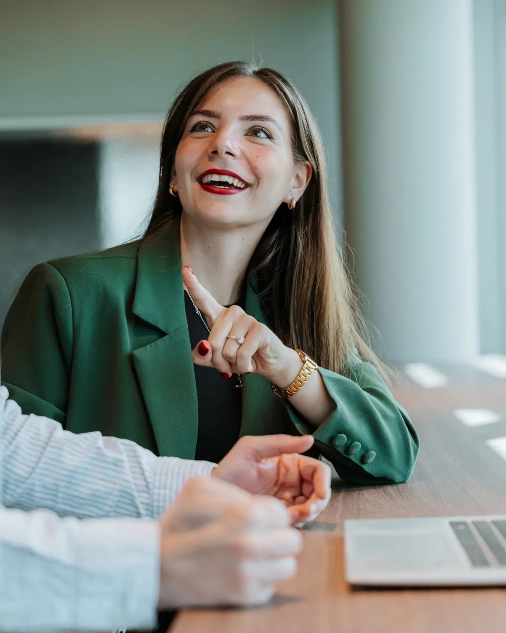 Woman in a green blazer smiling and gesturing while sitting at a table with a laptop, engaged in conversation.