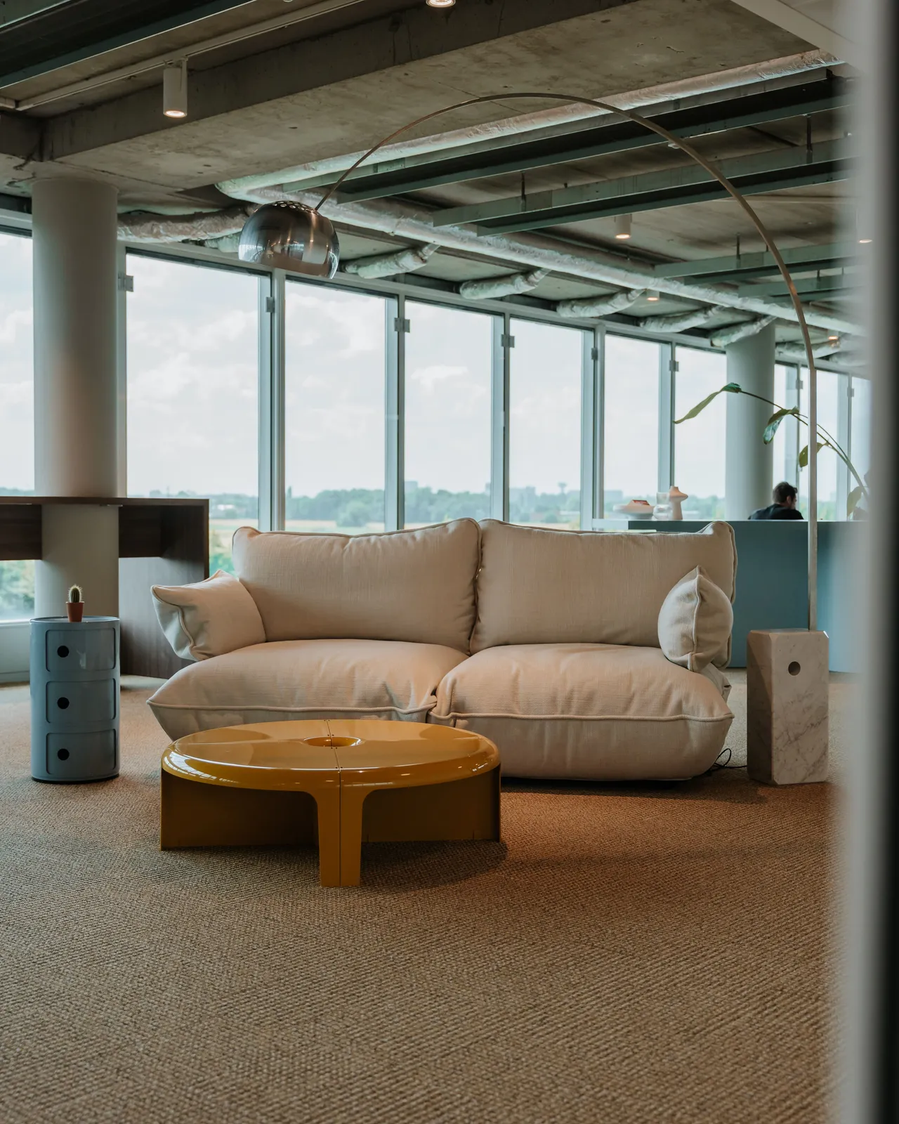 Modern lounge with a beige sofa, yellow coffee table, and large windows offering a cityscape view. Industrial ceiling and minimalist decor.