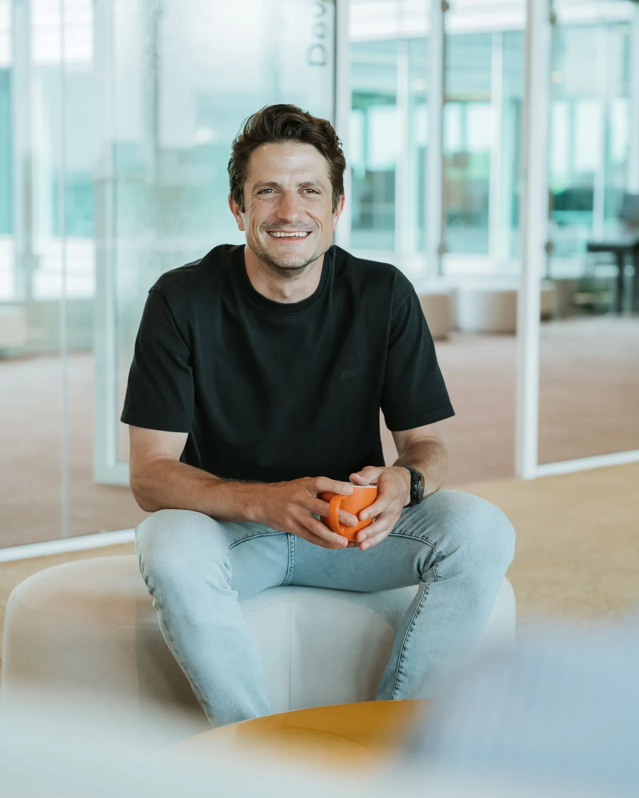 Smiling man in a black t-shirt and jeans sits on a white chair, holding an orange ball in a bright, modern office space.